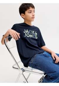 Αγόρι με κοντά σκούρα μαλλιά που φοράει μπλε μπλουζάκι "Jack & Jones" και μπλε τζιν, καθισμένο πλευρικά σε διαφανή καρέκλα με φόντο λευκό.