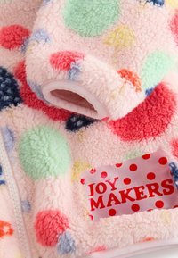 Fleecejacka med rosa bas, mångfärgade prickar och röd "JOY MAKERS"-etikett på rosa bakgrund; mjukt, strukturerat tyg.