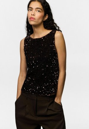 Femme aux longs cheveux bouclés portant un haut noir à sequins sans manches et un pantalon sombre à fines rayures, les mains dans les poches devant un fond uni.