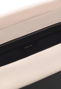 Wnętrze torby tote w kolorze czarnym i beżowym o gładkiej fakturze, z zamykaną kieszenią i wytłoczonym logo "TOUS". Charakteryzuje się kontrastowym przeszyciem.
