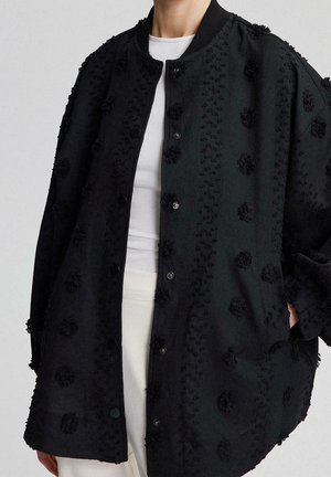 Zwarte oversized jas met getextureerde bloemenpatronen, open gedragen over een witte top en crèmekleurige broek, met zichtbare voor- en zijzakdetail.