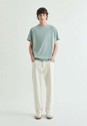 Joven de pie con las manos en los bolsillos, vestido con camiseta verde claro, pantalones blanco hueso y zapatillas blancas, sobre un fondo blanco liso.