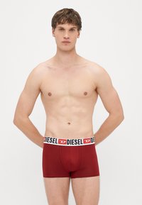 Červené bavlnené boxerky s čierno-bielym pásom s logom značky. Model stojí s neutrálnym výrazom, jasne prezentujúc výrobok.