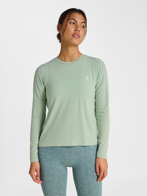 Vrouw draagt een lichtgroen shirt met lange mouwen en grijze getextureerde leggings, staande tegen een effen witte achtergrond, kijkt opzij.