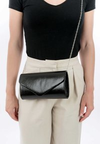 Femme portant un pantalon beige taille haute et un haut noir avec un petit sac bandoulière en cuir noir texturé, à chaîne argentée.