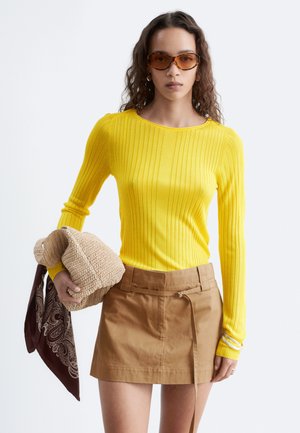 Femme aux cheveux bouclés portant un haut jaune côtelé à manches longues, une mini-jupe marron, des lunettes de soleil, tenant un sac tricoté beige avec une écharpe marron à motifs.