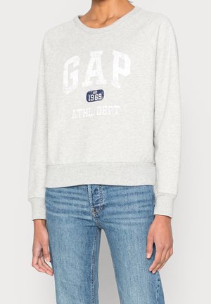 Person wearing light gray GAP sweatshirt with "EST. 1969 ATHL DEPT" text, paired with blue denim jeans against white background.