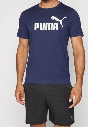 Mężczyzna w granatowej koszulce Puma z białym logo i czarnych szortach Puma stoi na jasnym tle.