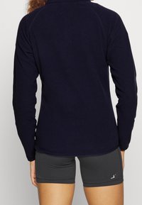 Marinblå fleecetröja med hög krage och lång ärm, mjuk textur, avslutad med en rundad nederkant. Parat med grå shorts.