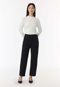 Top bianco a maniche lunghe abbinato a pantaloni neri a vita alta e a gamba larga. I pantaloni presentano una parte frontale liscia e spacco laterale. Tacchi a punta neri.
