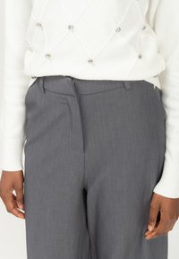 Mujer vistiendo un suéter blanco con acentos de perlas y pantalones de tiro alto grises, con las manos relajadas a los lados.