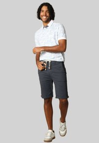 Korte mouwen, lichtblauw polo met palmbladvprint, gecombineerd met donkergrijze shorts met een trekkoord in de taille en opgerolde manchetten, gedragen met witte sneakers.