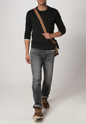 Hombre con suéter negro texturizado, jeans grises y botas de cuero marrón, llevando una bolsa mensajero color marrón claro, caminando frente a un fondo liso.