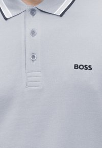 Polo gris clair à trois boutons, col rayé noir et blanc, avec le logo "BOSS" brodé sur la poitrine.