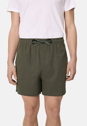 Hombre vistiendo pantalones cortos verdes olivo con cordón y bolsillos laterales, y una camiseta blanca, de pie frente a un fondo liso.