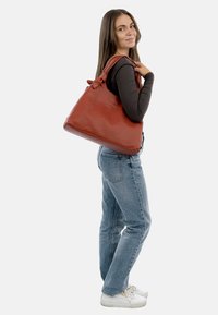 Bolso de cuero naranja con un acabado texturizado, de gran tamaño y con acentos de tira anudada, llevado al hombro por un modelo que viste ropa casual.