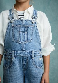 Denim tuinbroeken met verstelbare banden, lichtblauwe kleur, voorzak en zilverkleurige knopen, gecombineerd met een wit overhemd eronder.