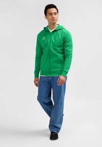 Grøn hættetrøje med lynlås og snøre i hætten, med et logo på brystet, kombineret med løse blå cowboybukser og sorte sko.