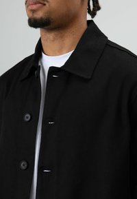 Zwarte jas van gestructureerde stof, met een kraag, twee knopen en minimale stikdetails. Gedragen over een wit overhemd.