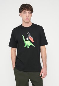 Μαύρο βαμβακερό μπλουζάκι με γραφικό πράσινου δεινοσαύρου, συνοδευόμενο από μπρελόκ και κόκκινο αξεσουάρ. Κοντά μανίκια και κλασική εφαρμογή.