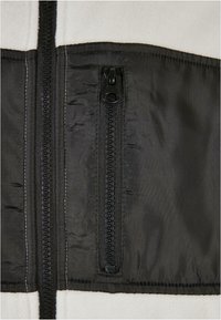 Gros plan d'une poche zippée noire sur un vêtement en tissu blanc et noir, montrant des coutures détaillées et un curseur de fermeture éclair.