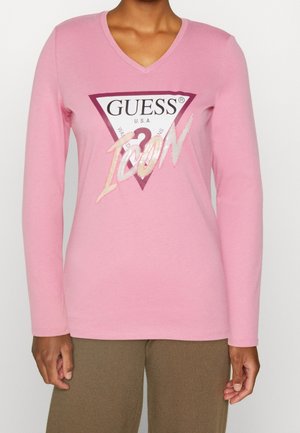 Personne portant un t-shirt rose à col en V manches longues avec un grand logo "GUESS Icon" et un pantalon marron, sur un fond uni.