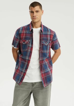 Mężczyzna w krótkim czerwono-niebieskim kratkę koszuli zapinanej na guziki, na białej koszulce, w zestawieniu z szarymi dżinsami, stoi z rękami za plecami.