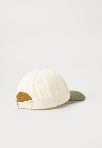 Baseball cap with cream-colored fabric crown and adjustable strap, dark olive green curved brim, set against plain white background.