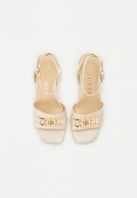 Beige slingback pete z visoko peto s kvadratnim nosom, sijočim paten finim zaključkom in zlatim okrasom na trakcu. Gladka tekstura, minimalističen dizajn.