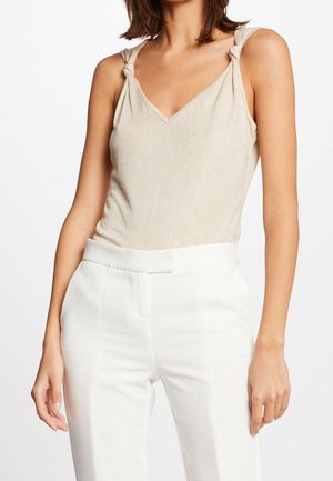 Mujer con un top beige sin mangas de escote en V con tirantes anudados y pantalones blancos de talle alto, posando frente a un fondo liso.
