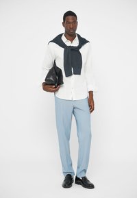 Chemise blanche à boutons, pantalon rayé bleu clair, pull marine foncé noué autour du cou, tenant un sac noir, chaussures noires.