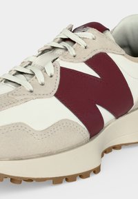 Gros plan d'une basket beige et blanche avec le logo "N" bordeaux, des lacets blancs et une semelle en gomme texturée sur fond uni.