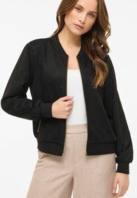 Juoda bombonešio striukė iš minkšto zomšos primenančios medžiagos. Turi auksinę užtrauktuką, dryžuotus rankogalius ir apačią, bei laisvo kirpimo. Derinama su gelsvais kelnėmis.