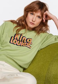 Felpa verde con grafica "King Louise" in un carattere arrotondato multicolore. Presenta un collo a giro e polsini a coste. Tessuto leggero e testurizzato.