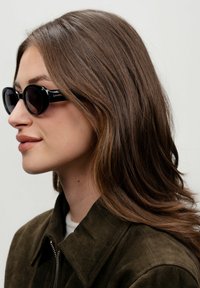 Černé sluneční brýle s kulatými rámem, lesklým povrchem a tónovanými skly. Na sobě má tmavozelenou semišovou bundu, která podtrhuje moderní design.