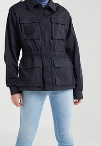 Chaqueta utilitaria azul marino fabricada en tejido ligero, con cuatro bolsillos frontales y un cinturón con cordón ajustable en la cintura.