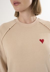 Felpa beige con collo a girocollo a coste, caratterizzata da un piccolo cuore rosso ricamato sul lato sinistro del petto e dettagli delle cuciture ben visibili.
