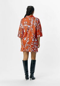 Vestido naranja con patrones florales blancos, mangas cortas, corte holgado y un collar; combinado con botas altas negras brillantes.