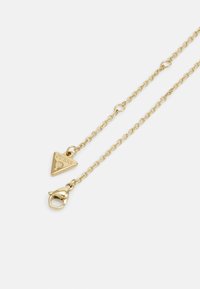 Collier chaîne en or avec un petit pendentif triangulaire gravé de "GUESS" et une fermeture à mousqueton sur fond blanc.