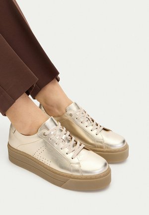 Piedi che indossano sneakers platform metalliche dorate con suole beige e pantaloni marrone chiaro su uno sfondo semplice.