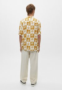 Camisa de manga corta en un patrón a cuadros amarillo y blanco con diseño de hojas de palmera. Lleva pantalones sueltos de color claro y zapatos negros.