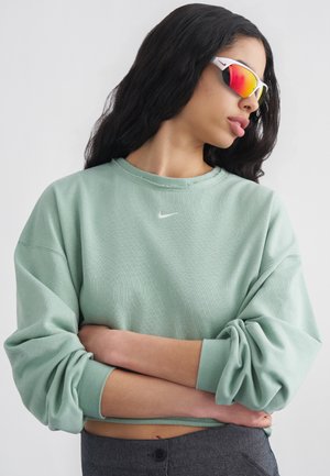 Kobieta o długich ciemnych włosach, nosząca refleksyjne czerwone sportowe okulary przeciwsłoneczne i jasnooliwianą bluzę Nike z założonymi rękami.