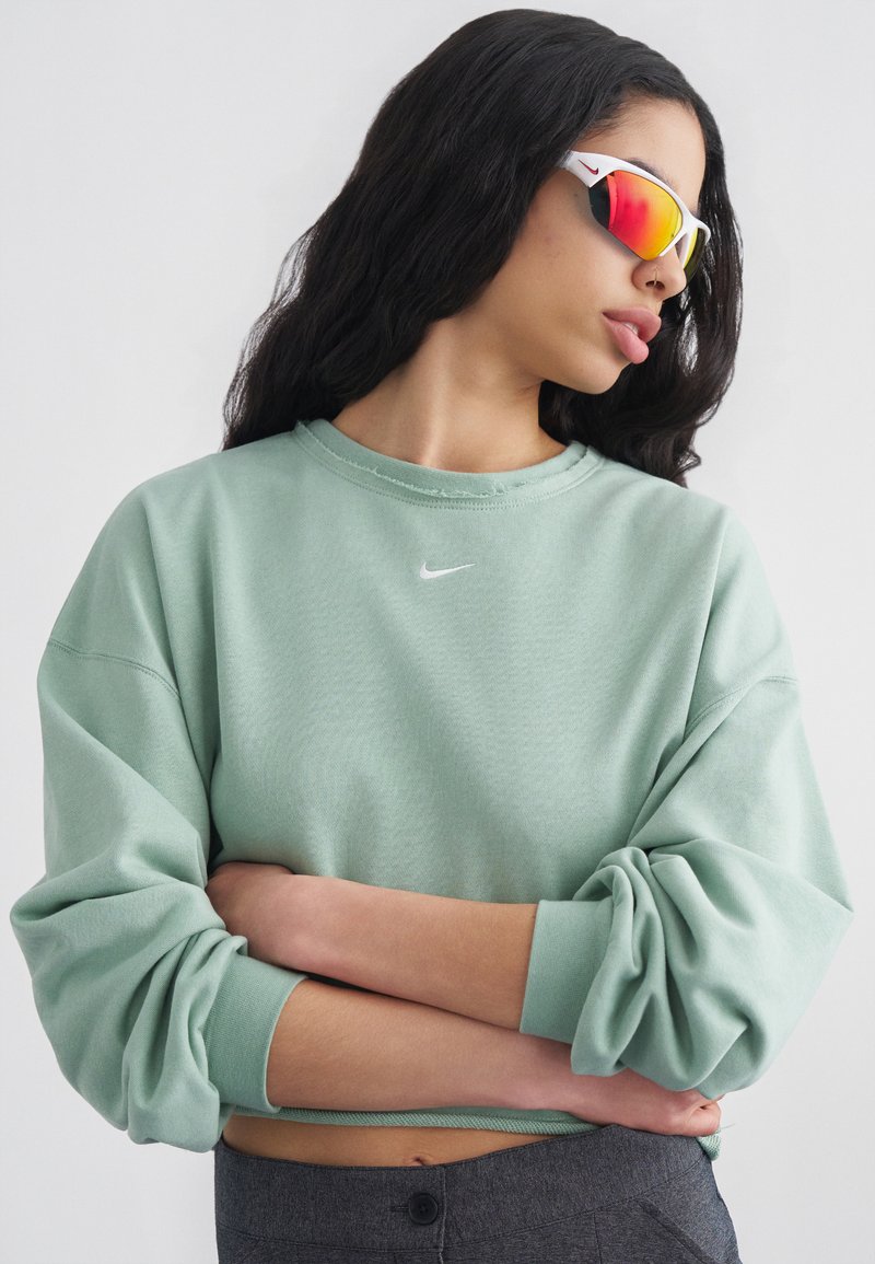 Γυναίκα με μακριά σκούρα μαλλιά που φορά αντανακλαστικά κόκκινα αθλητικά γυαλιά ηλίου και ένα ανοιχτοπράσινο φούτερ Nike με τα χέρια σταυρωμένα.