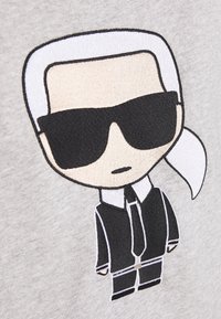 Camiseta gris con un personaje de dibujos animados bordado, que tiene el cabello blanco, gafas de sol negras y un traje negro, que incluye una corbata blanca y un pañuelo de bolsillo.