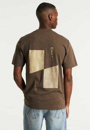 Camiseta de algodón marrón que presenta dos cuadrados texturizados de color marrón claro con un patrón topográfico y texto vertical en blanco en la parte posterior.