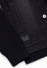 Gros plan d'une veste en denim noire avec manchette en maille côtelée, couture visible de la poche avant et bouton-pression en métal avec texte embossé.
