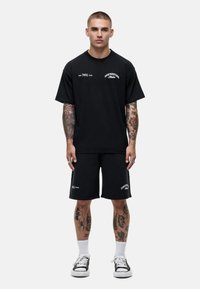 Zwarte katoenen graphic t-shirt en bijpassende shorts, met witte tekst op de voorkant. Model draagt zwarte sneakers en witte crew sokken.