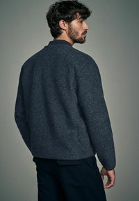 Grijze wollen jas met een eenvoudig ontwerp, voorzien van een ronde kraag en lange mouwen, gecombineerd met donkere broeken. Gladde textuur, minimale details.