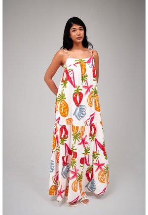 Kobieta w białej sukience maxi z kolorowymi nadrukami owoców i rozgwiazd, stojąca ze splecionymi rękami za plecami na gładkim jasnoszarym tle.