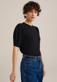 Svart kortärmad topp med puffärmar, rund halsringning, tillsammans med högmidjade blå jeans och ett smalt svart bälte. Slät textur.